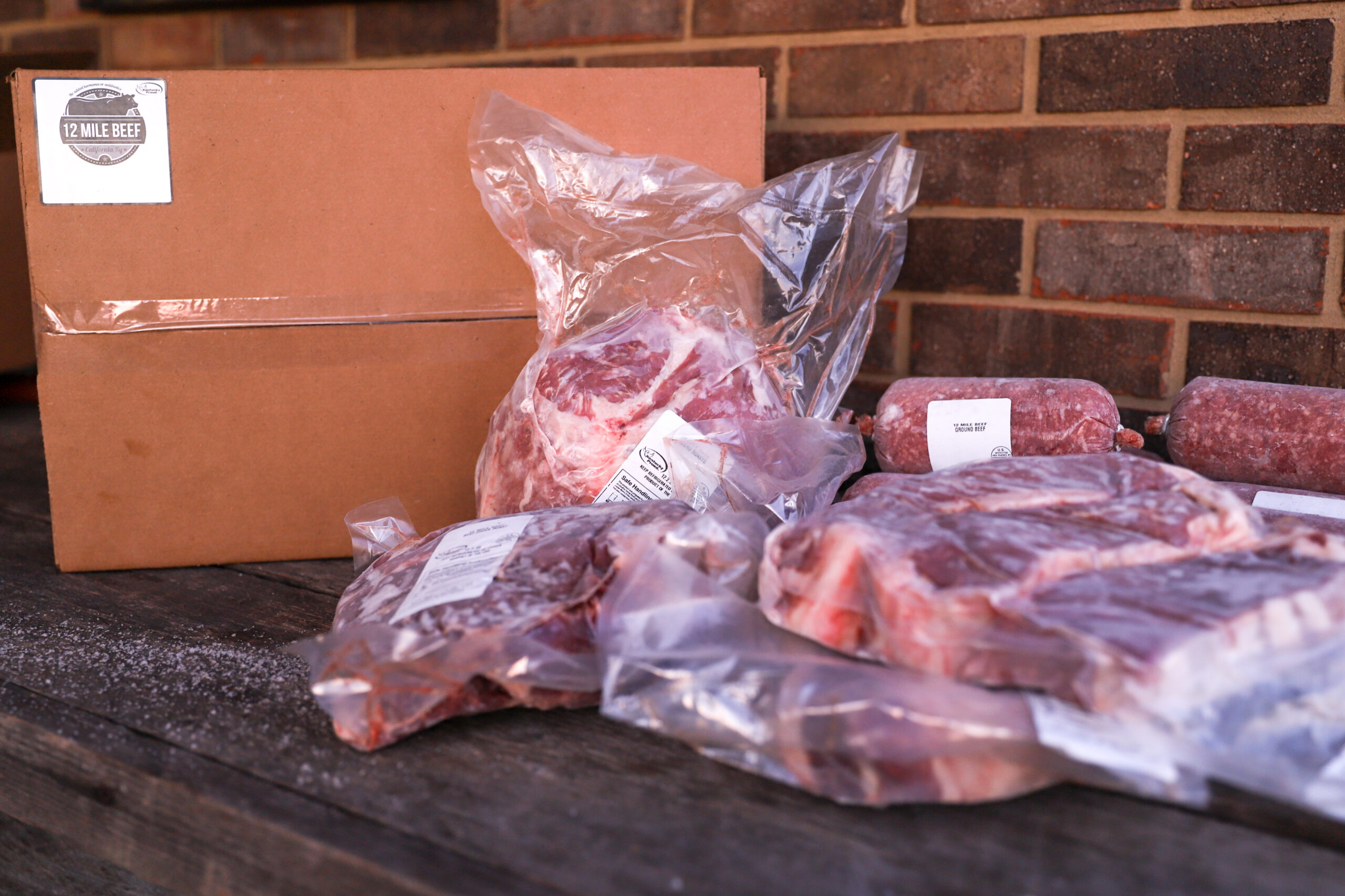 Assorted cuts of beef from 12 Mile Beef, including packaged steaks and ground beef, displayed alongside a cardboard delivery box on a wooden surface.
