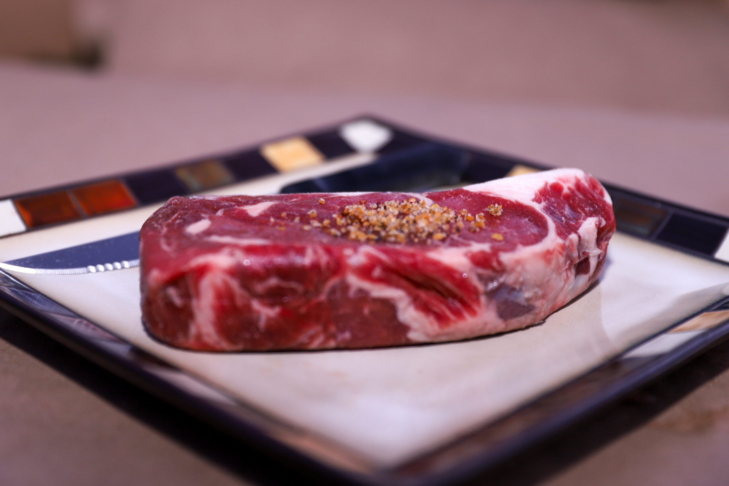 Raw ribeye steak seasoned with spices on a decorative plate, ready for cooking.
