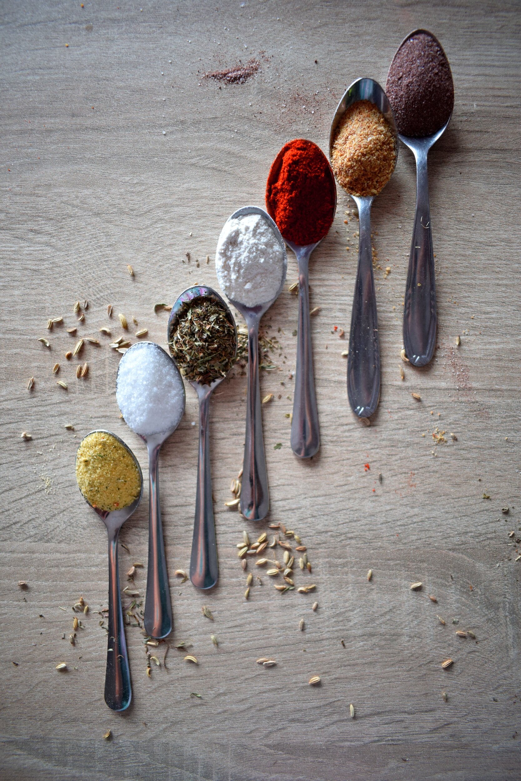 Assorted spices in silver spoons arranged on a wooden surface, including paprika, salt, and dried herbs, with scattered seeds and spice dust in the background.