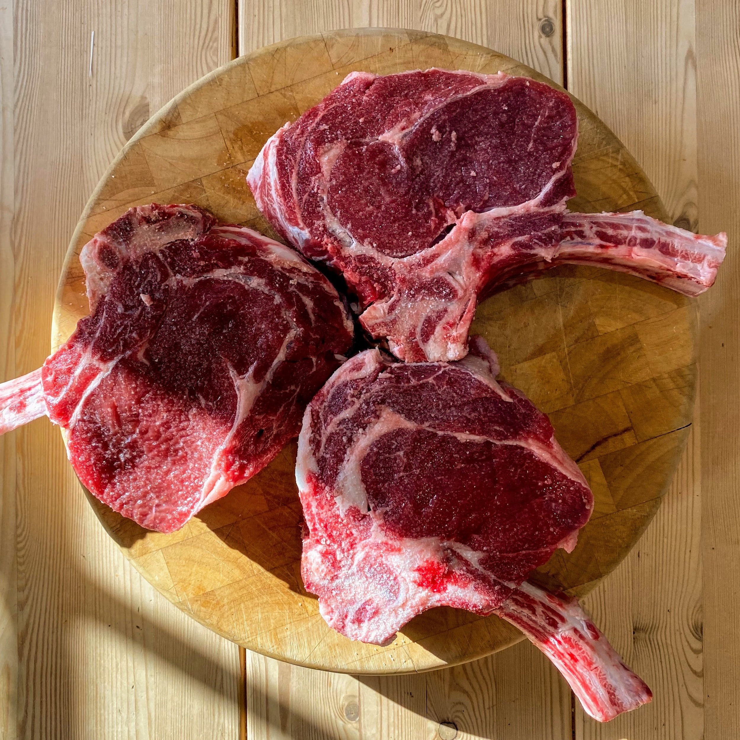 unsplash-image-6-5Ul3I6vSE Three raw tomahawk steaks resting on a wooden cutting board, showcasing their marbled texture and thick cut, ideal for grilling or roasting.
