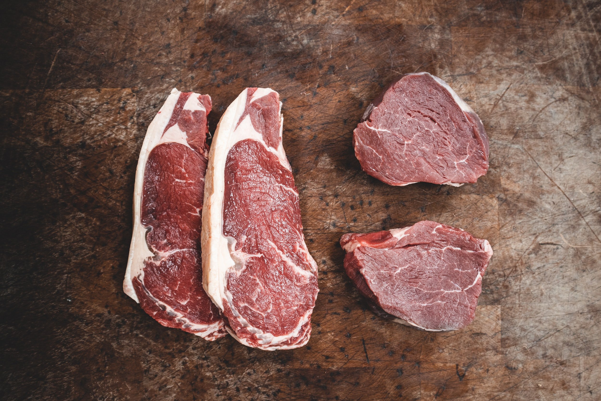 unsplash-image-lvmFFcLvHl8 Fresh cuts of beef steak displayed on a wooden cutting board, featuring two ribeye steaks and two smaller sirloin cuts, showcasing marbling and rich red color ideal for grilling or roasting.